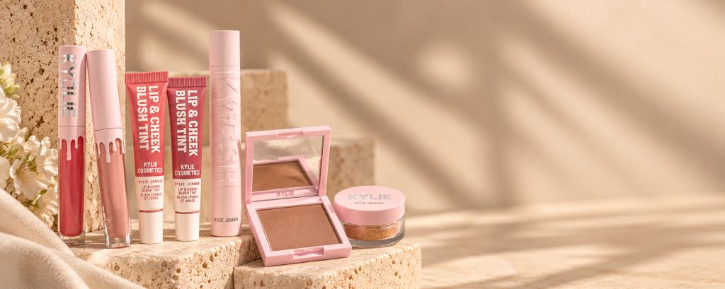 Kylie Cosmetics products in pink packaging: lip glosses, lip and cheek blush tints, mascara, a bronzer compact, and setting powder.