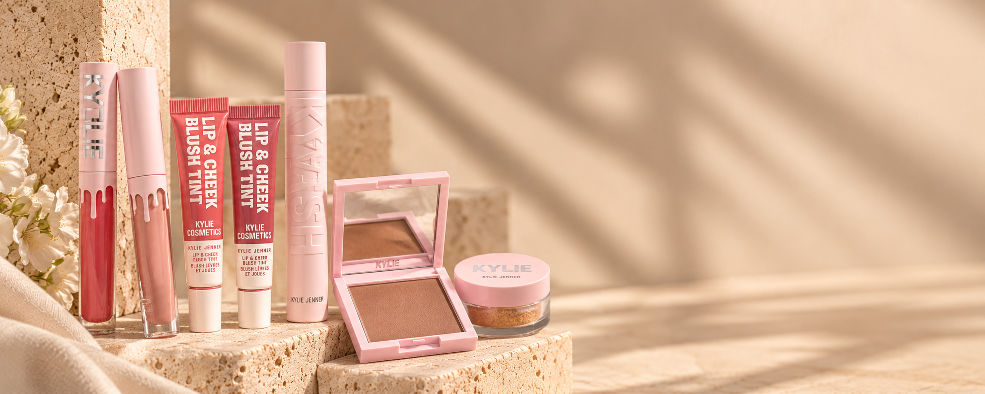 Kylie Cosmetics products in pink packaging: lip glosses, lip and cheek blush tints, mascara, a bronzer compact, and setting powder.