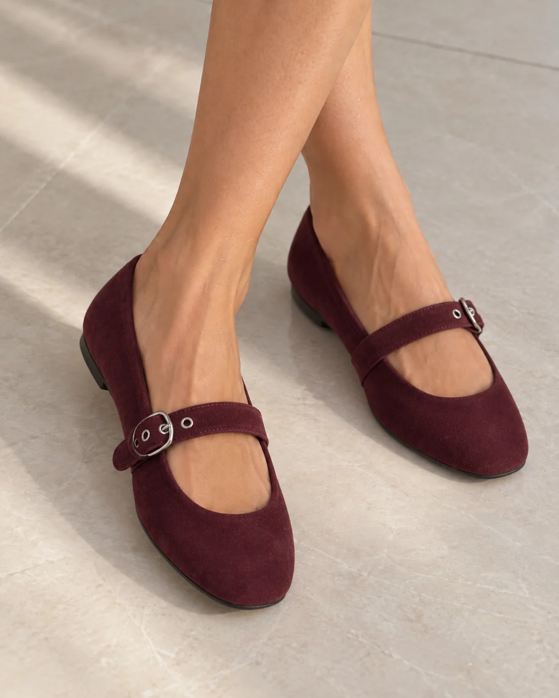 Burgundy suede Mary Jane flats with a buckled strap, worn on pale stone