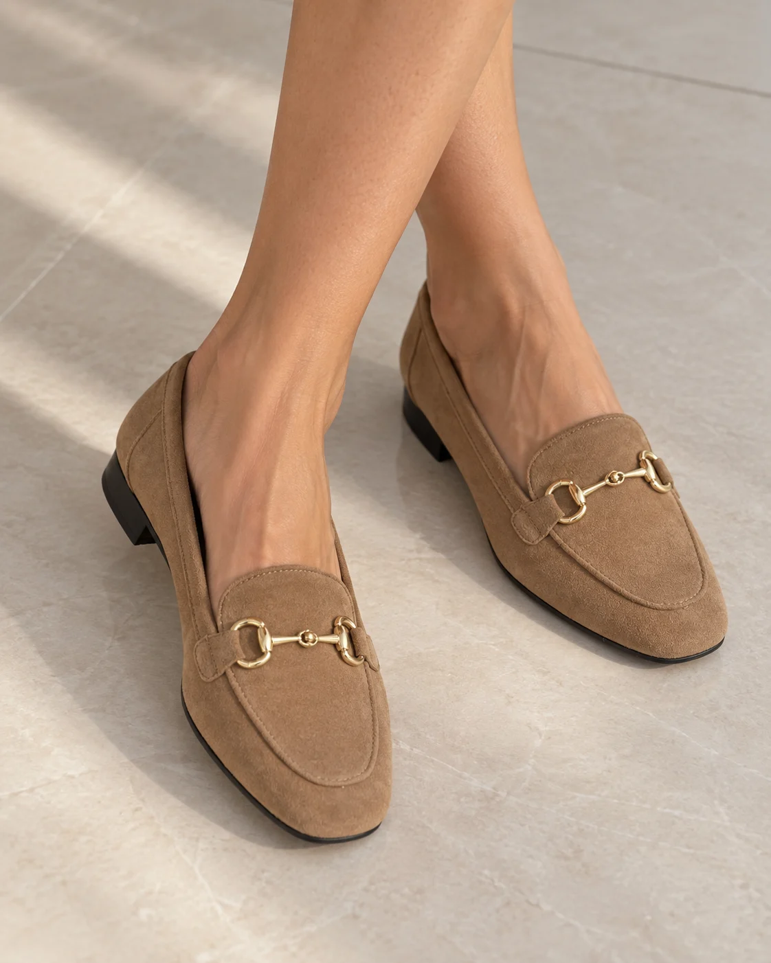 Camel suede loafers with a gold snaffle trim, worn on pale stone flooring