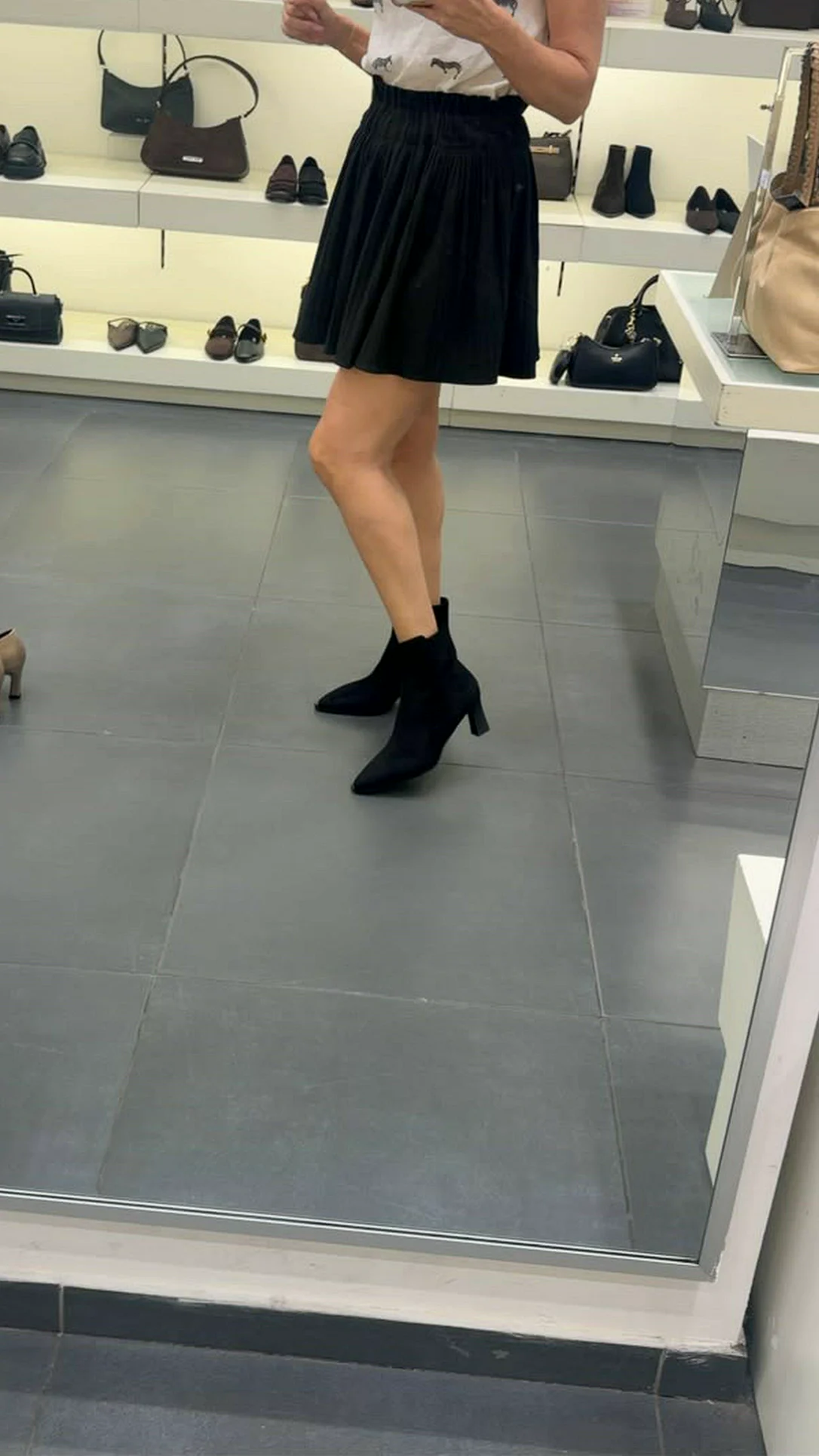 מיכל לורן במראה בחנות Nine West עם חצאית שחורה ומגפונים שחורים