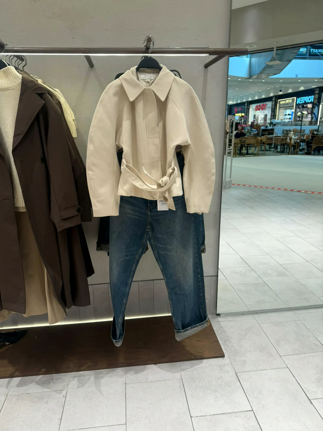 ג׳קט שמנת עם קשירה ומעיל חום על מתלה בחנות Zara